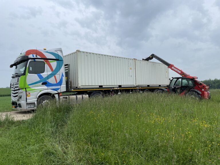 Camion transportant deux containers de stockage blancs sur un terrain herbeux, avec une grue Manitou manipulant l'un des containers.