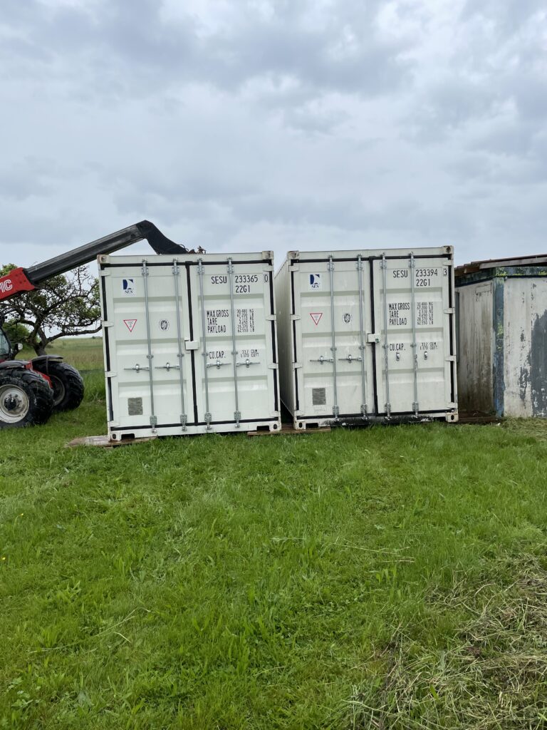 Deux containers de stockage blancs installés sur un terrain herbeux, avec une grue Manitou visible à gauche.