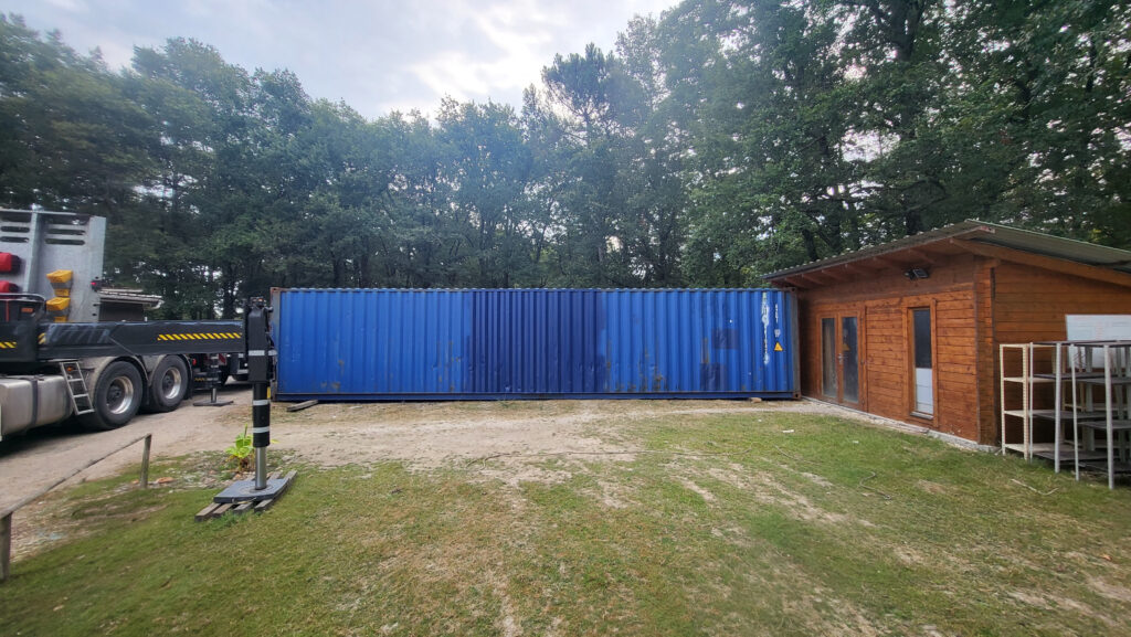 Container 40 pieds d'occasion bleu livré par In'Box Container chez NICO BRICO 24, à côté d'un bâtiment en bois, avec un camion de livraison à gauche.