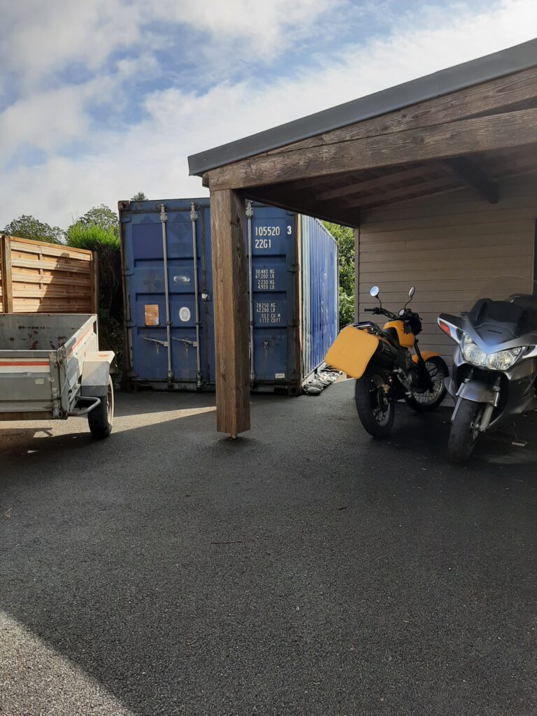 Un container maritime bleu de 20 pieds installé dans une cour, utilisé comme abri de jardin pour ranger une remorque, une moto jaune et un scooter.