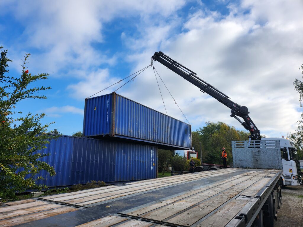 Un container maritime bleu de 40 pieds en cours de levage par une grue, avec un autre container déjà positionné en dessous. Un camion et des ouvriers sont visibles sur le site, sous un ciel partiellement nuageux.