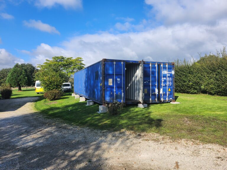 Deux containers maritimes bleus de 40 pieds installés côte à côte sur un terrain verdoyant, soutenus par des blocs en béton, avec des véhicules à l'arrière-plan sous un ciel dégagé.