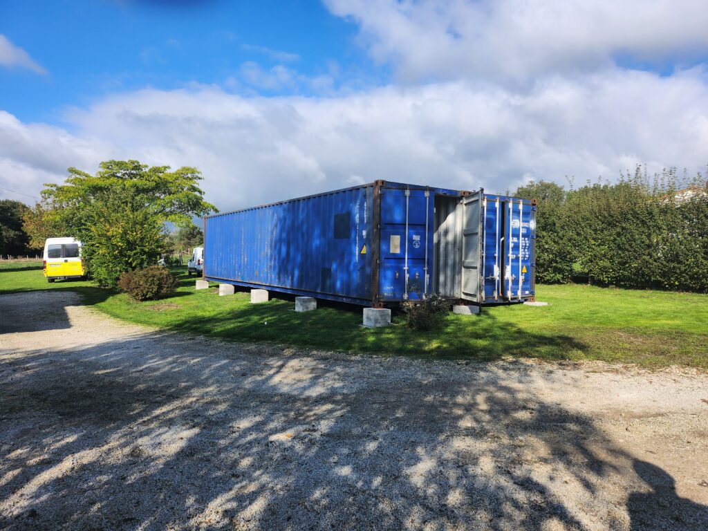 Deux containers maritimes bleus de 40 pieds positionnés côte à côte sur un terrain, reposant sur des blocs en béton. Un véhicule et des arbres sont visibles en arrière-plan sous un ciel dégagé.