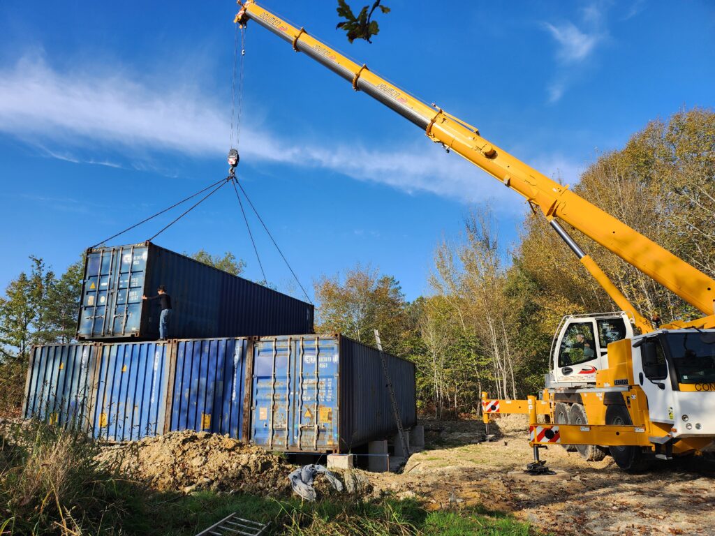 Une grue jaune place un container 40 pieds HC sur une pile de containers dans un chantier à Cornille, Dordogne. Un ouvrier est positionné sur le dessus pour guider l’installation.