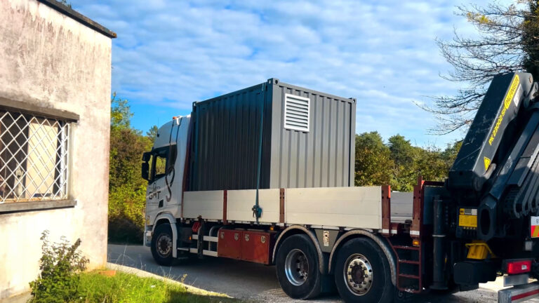 Un container gris de 10 pieds équipé d'une grille de ventilation, transporté sur un camion équipé d'une grue de la société GHT, dans un environnement arboré.