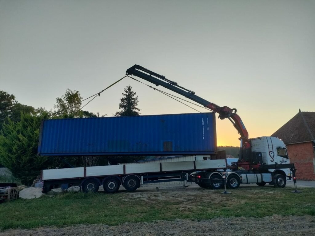 Un container maritime bleu de 40 pieds en cours de déchargement à l'aide d'une grue montée sur un camion, sous un ciel au coucher de soleil.