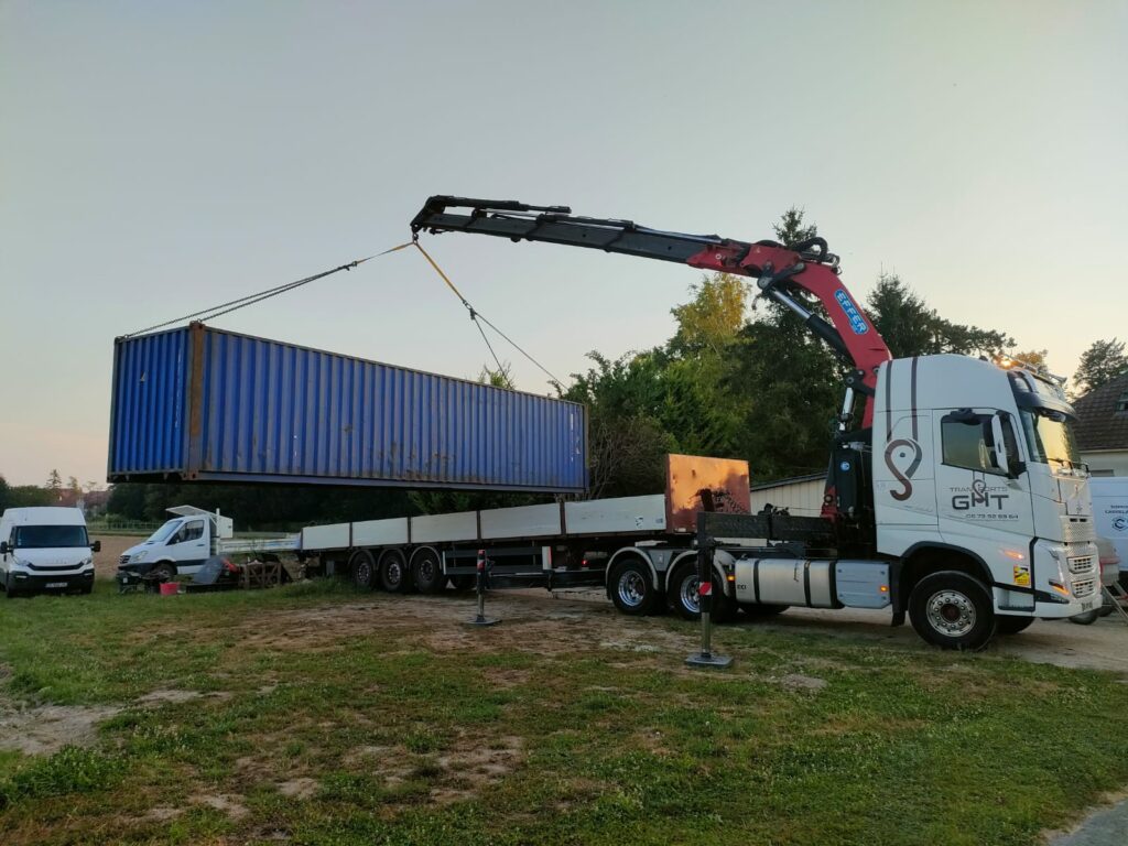 Un container maritime bleu de 40 pieds suspendu en l'air par une grue montée sur un camion lors d'une opération de déchargement sur un terrain.