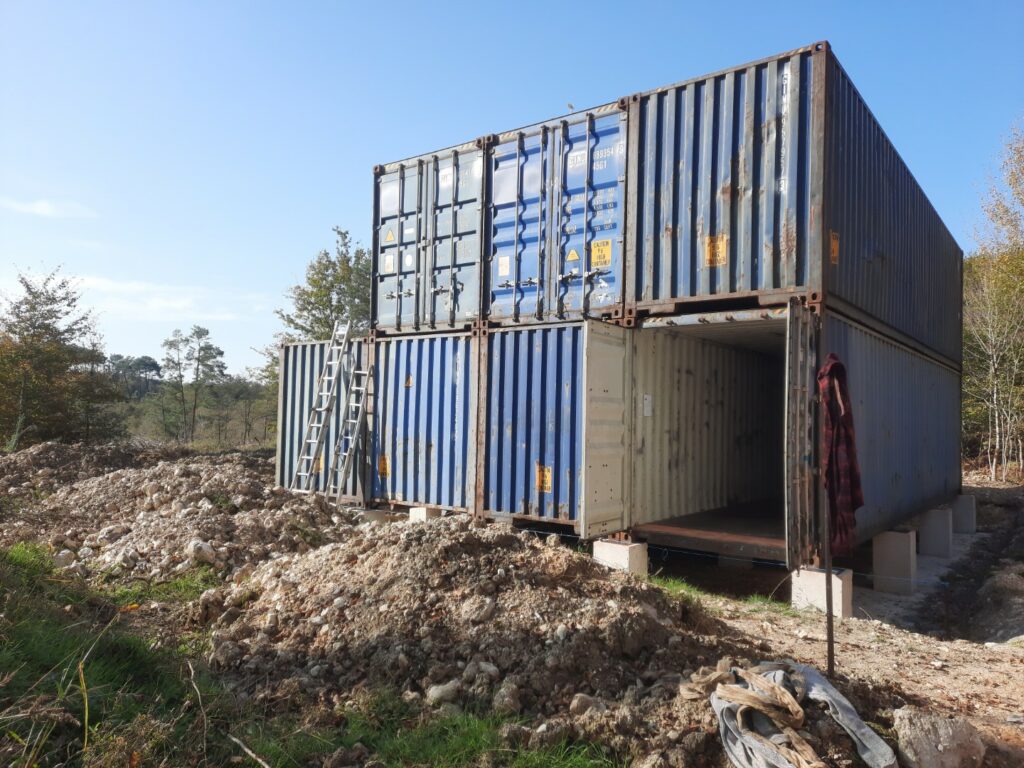 Sept containers 40 pieds HC empilés, partiellement aménagés, installés sur des blocs de béton dans un terrain de construction à Cornille, Dordogne.